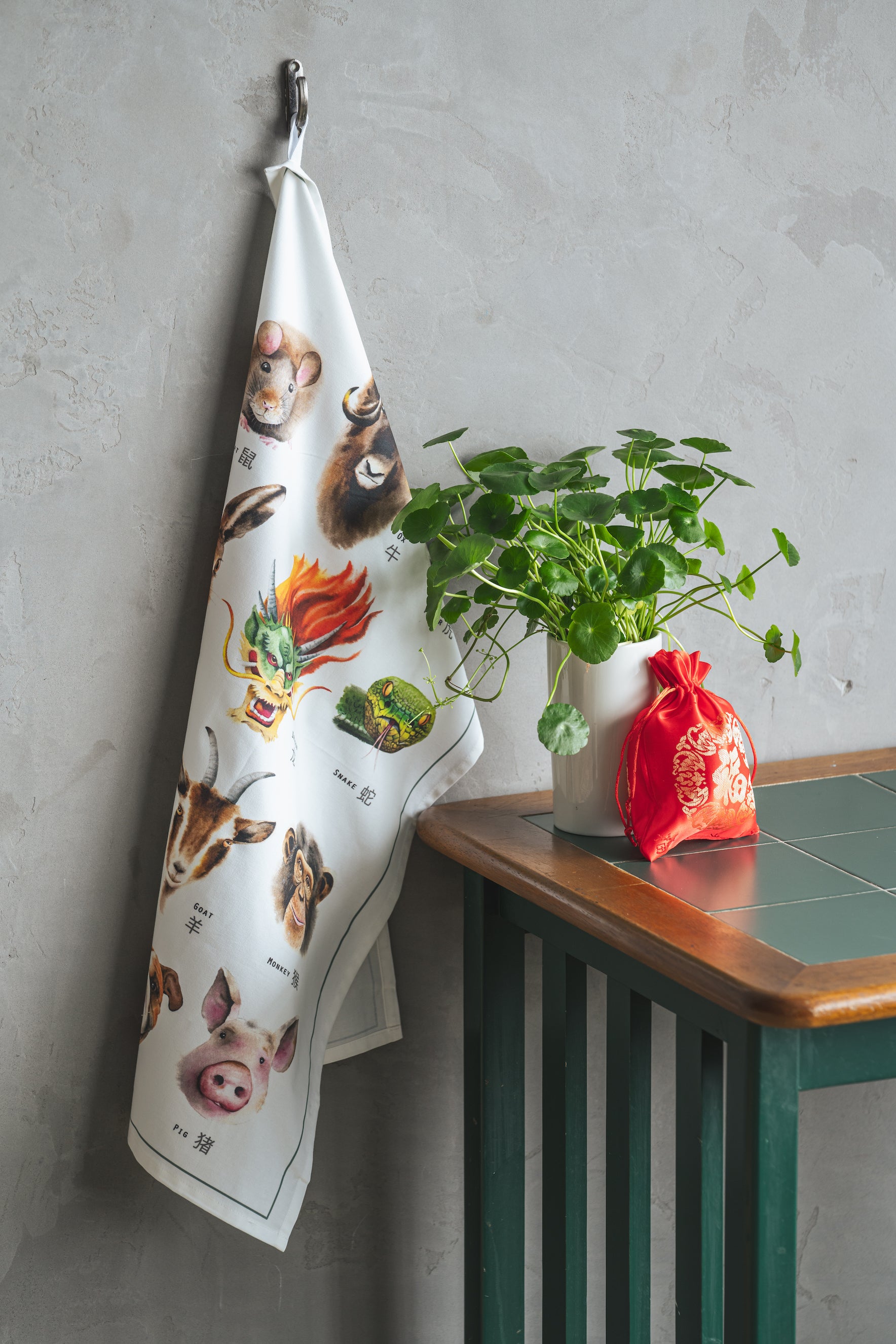 Lifestyle image of zodiac tea towel against a table and plants with a red bag.