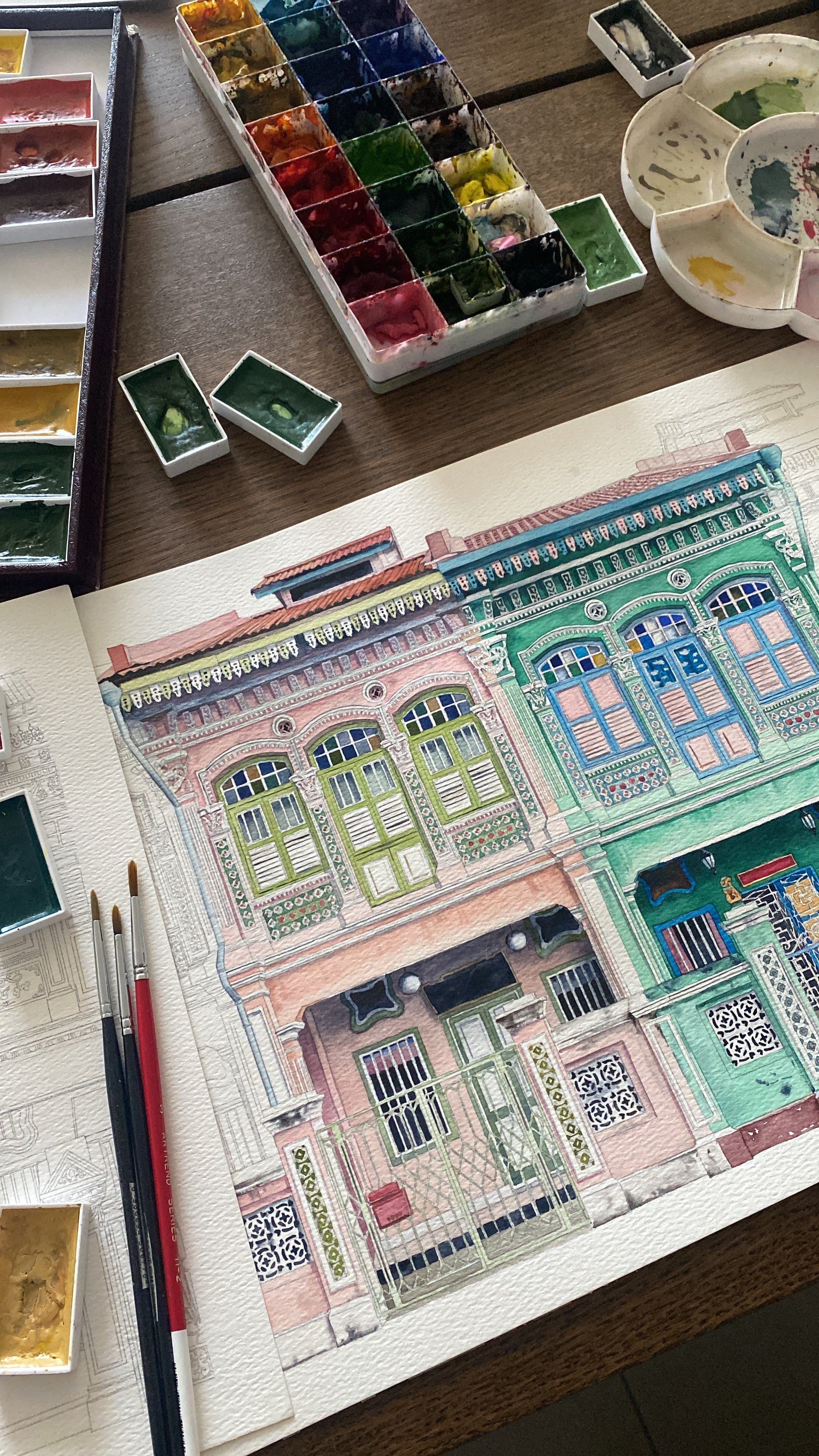 Artistic depiction of peranakan shophouses with buildings, watercolor palette, and brushes on a wooden surface.