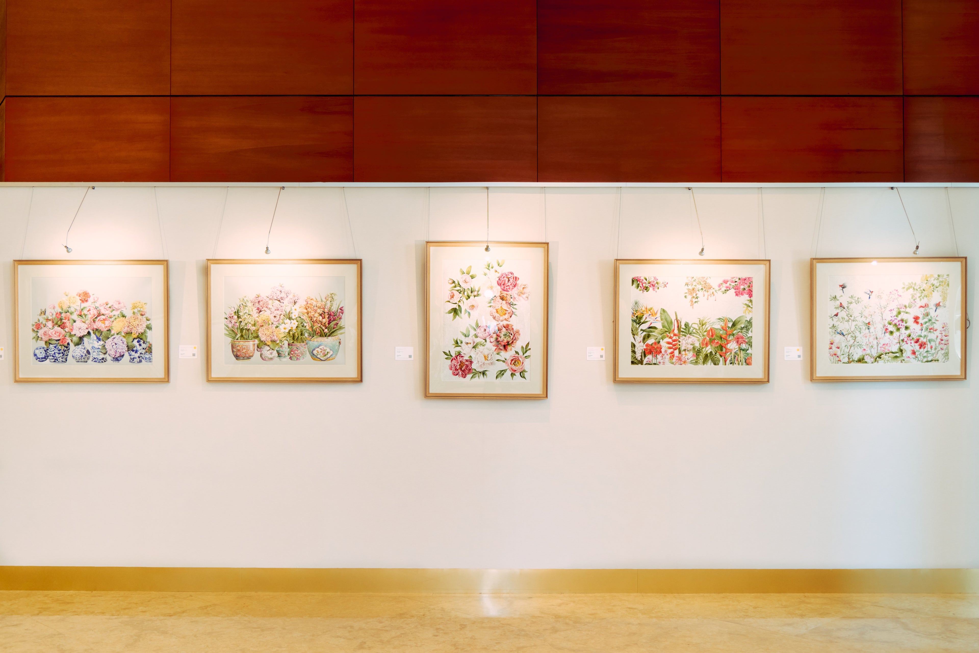 Five framed floral paintings on a white wall with a wooden floor and paneling.