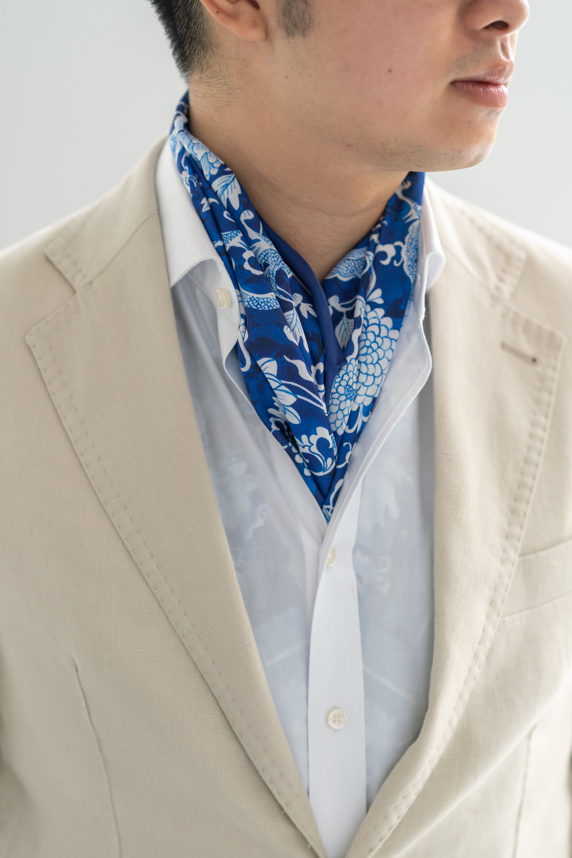A person wearing a beige blazer and a white shirt, with a blue silk scarf featuring a floral and dragon pattern around the neck.