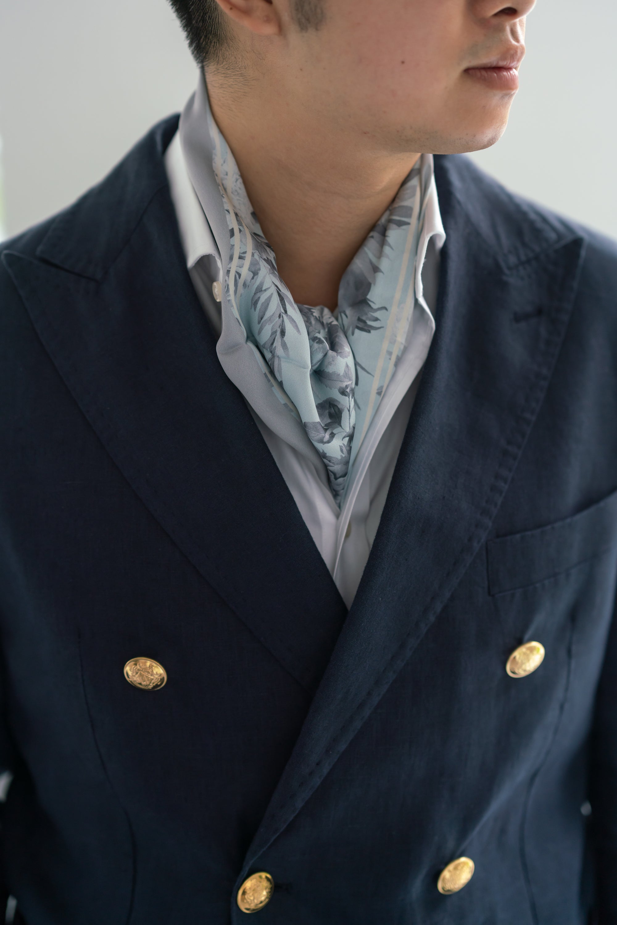A person wearing a grey floral silk scarf tied around the neck, with a navy blazer and a button-up shirt.