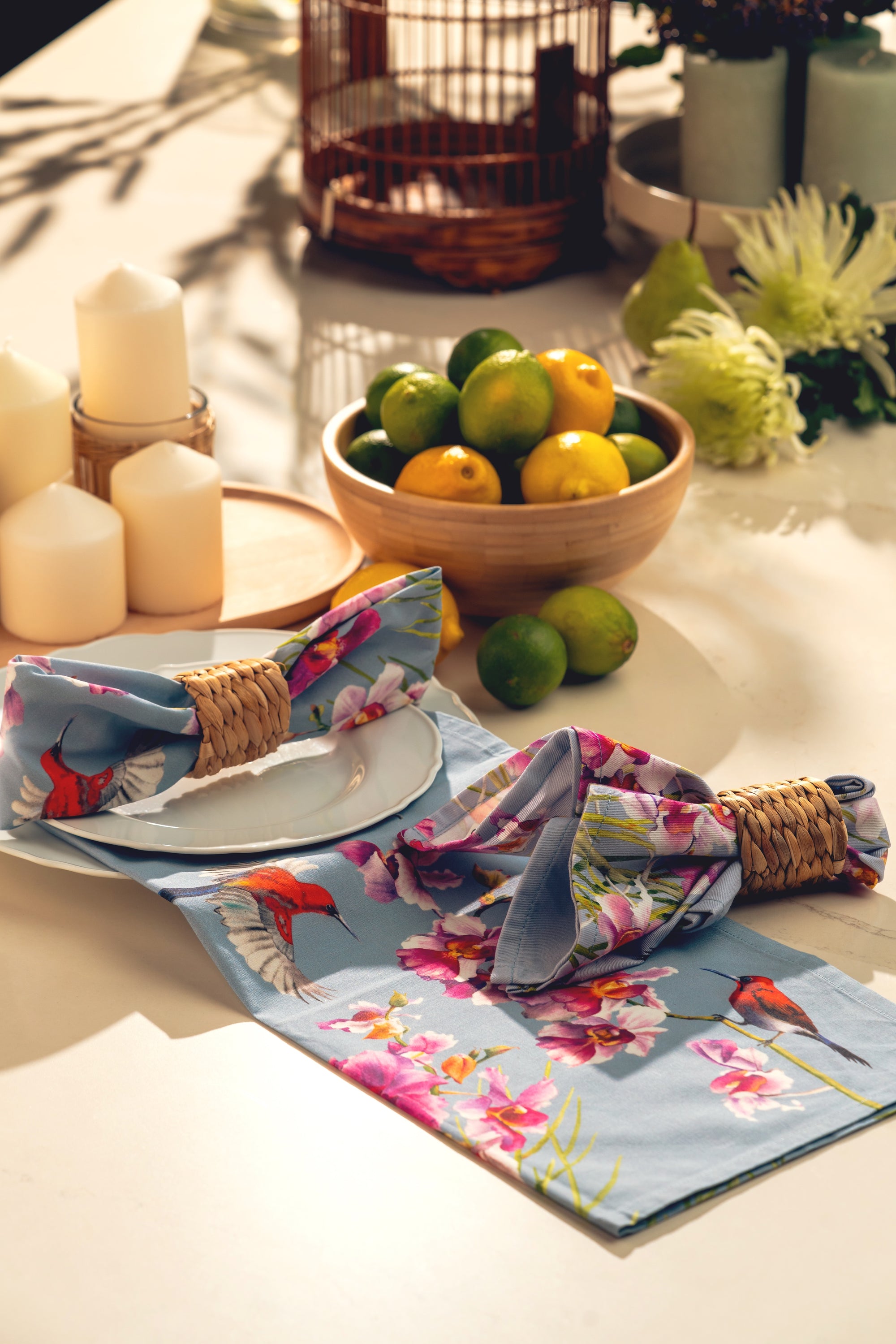 Decorative table setting with floral napkins, candles, and a bowl of fruit on a white surface.