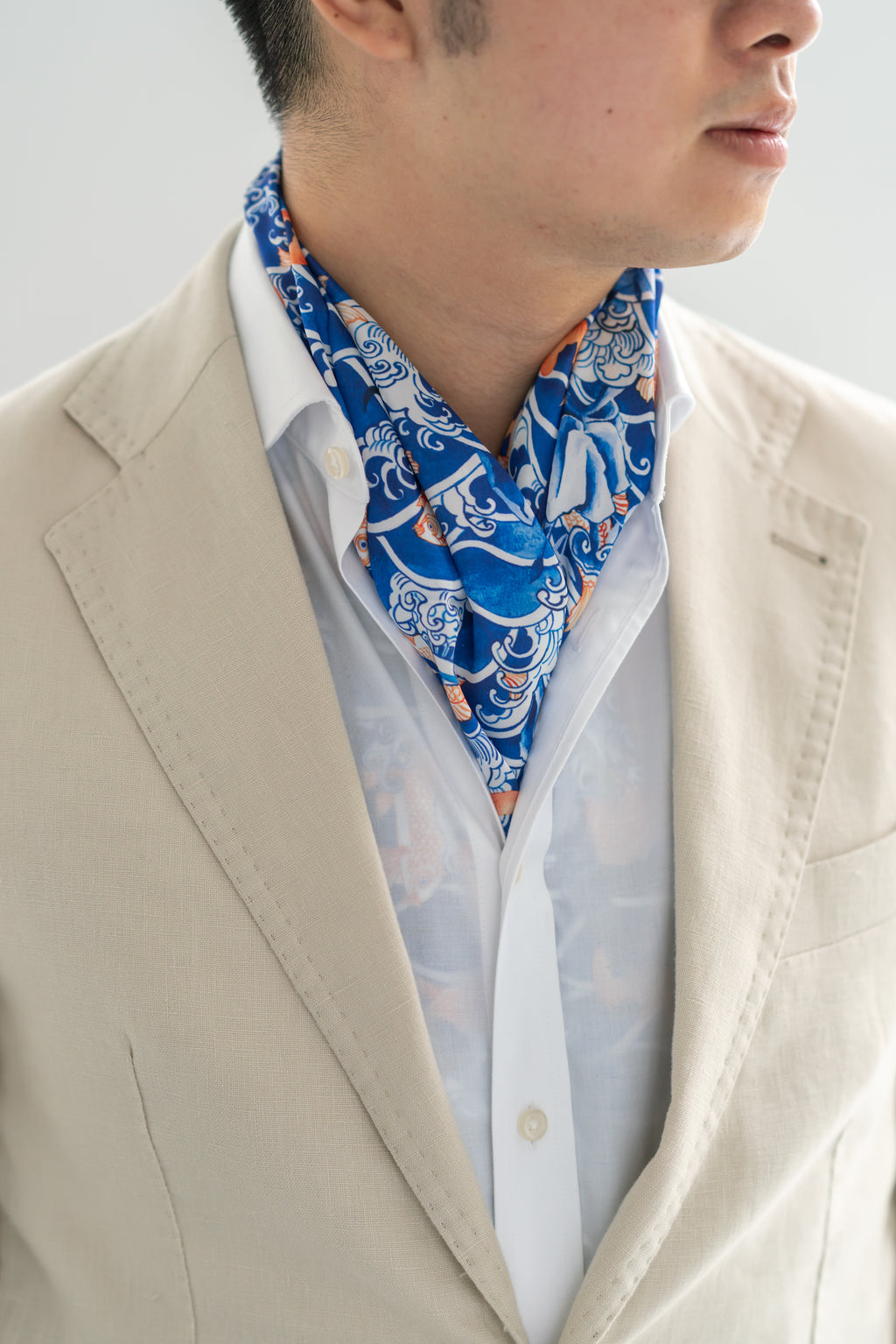 A person wearing a beige blazer and a white shirt, with a blue silk scarf featuring a wave pattern tied around the neck.