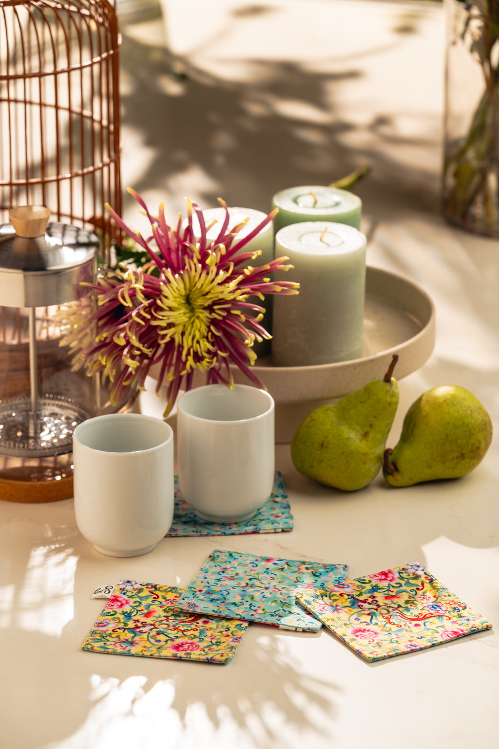 Ceramic cups with colorful coasters on a table with pears and a candle.