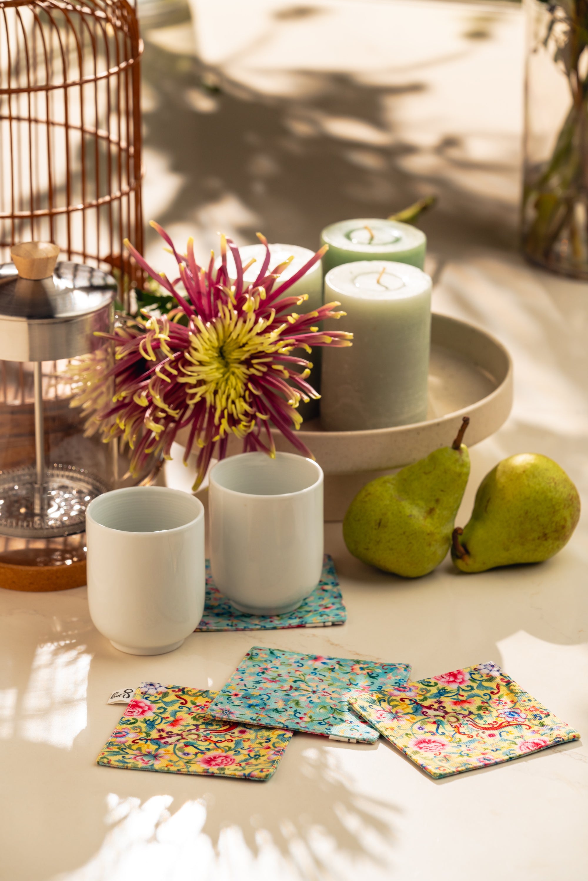 Ceramic cups with colorful coasters on a table with pears and a candle.