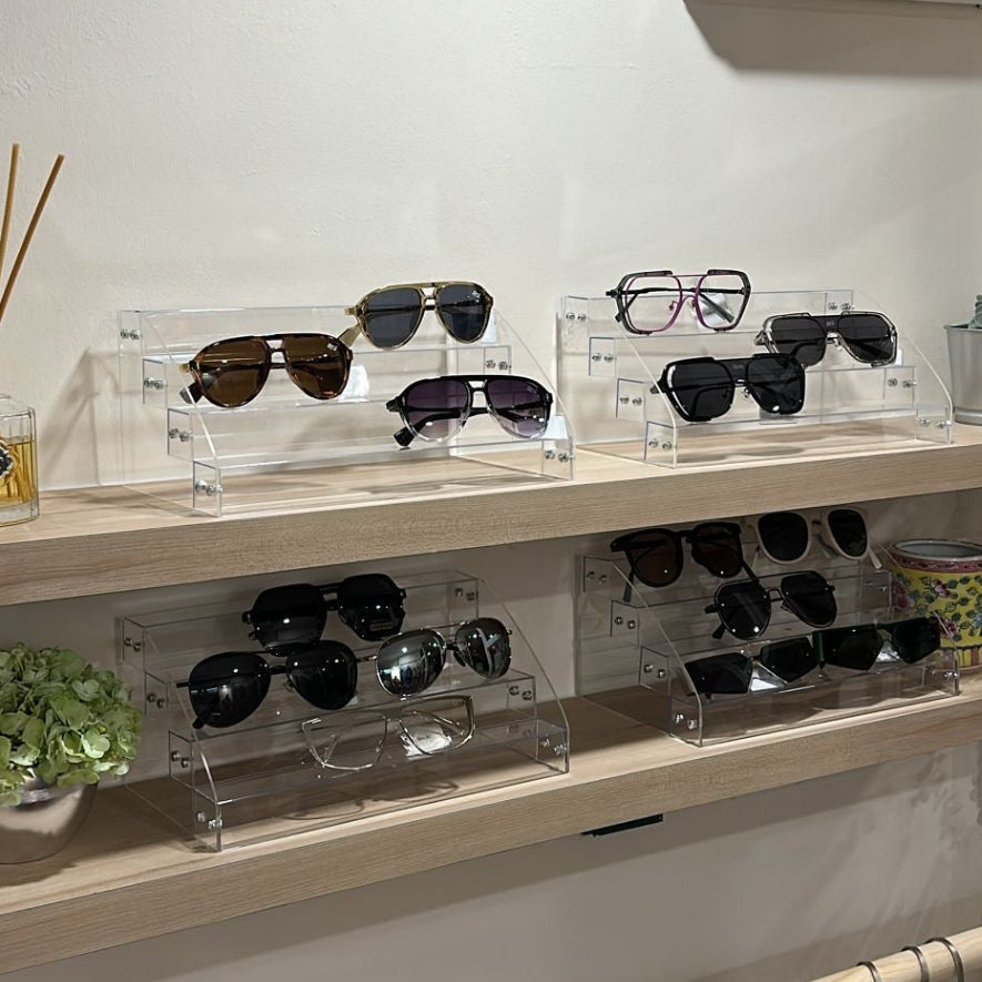 Display of sunglasses on a shelf with clothing racks below in a store setting.